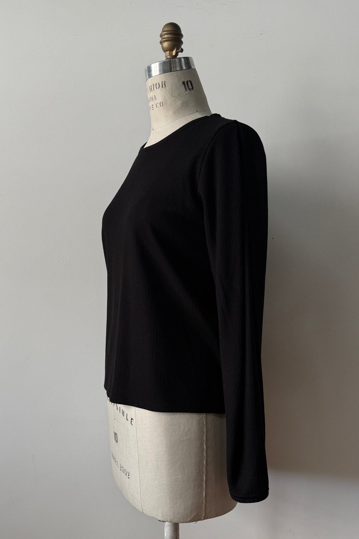 The Virginia Top by Kollontai in Black, a corduroy-textured top with inverted pleats on the long sleeves, a slim fit and a round neck, is shown on a mannequin against a grey background. 