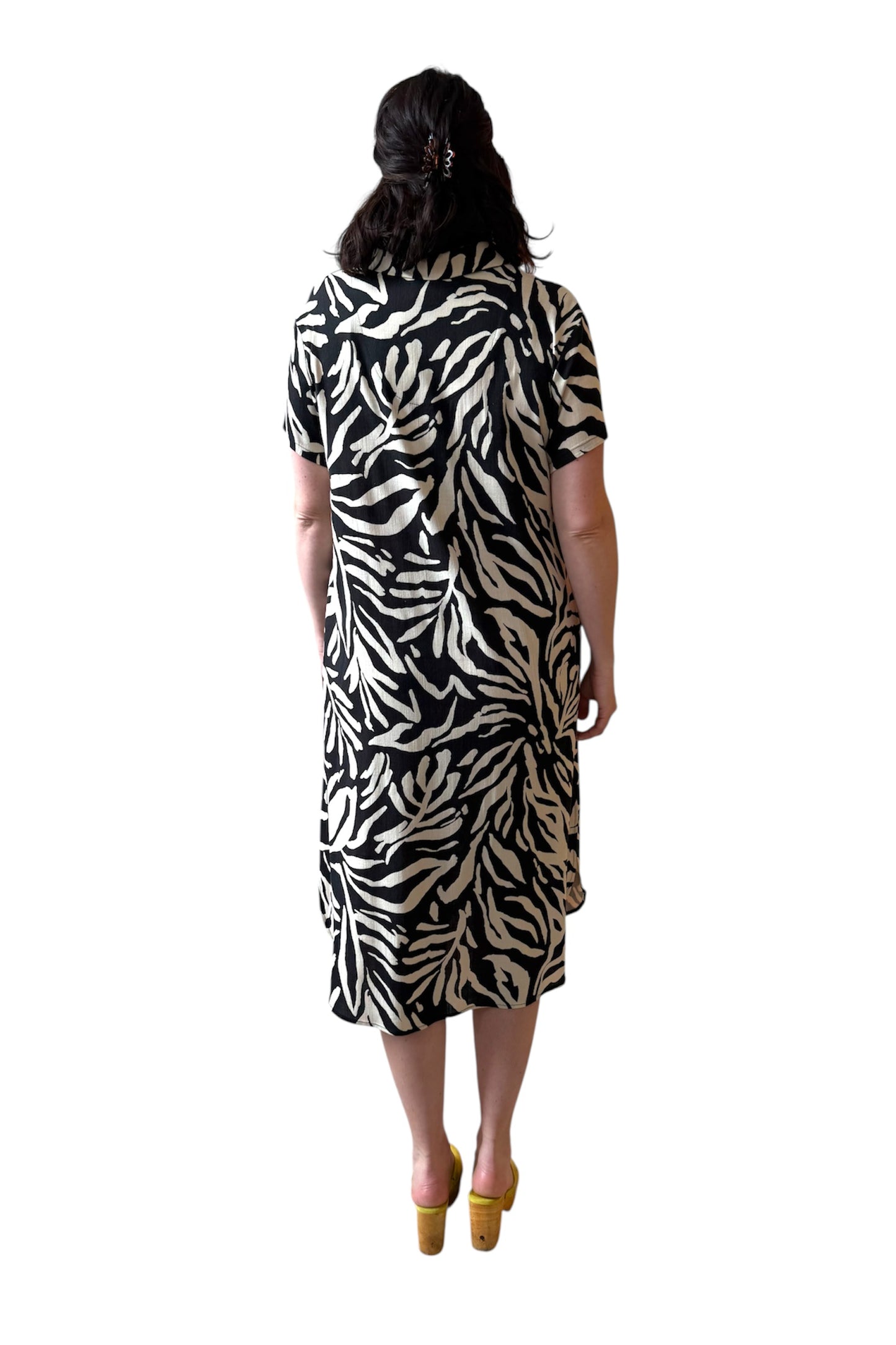 Back view of a woman wearing the Regina Dress by Compli K in Black and White print, a long dress with a v placket neck, short sleeves, loose fit, rounded hem and side slits. She is standing against a white background. 