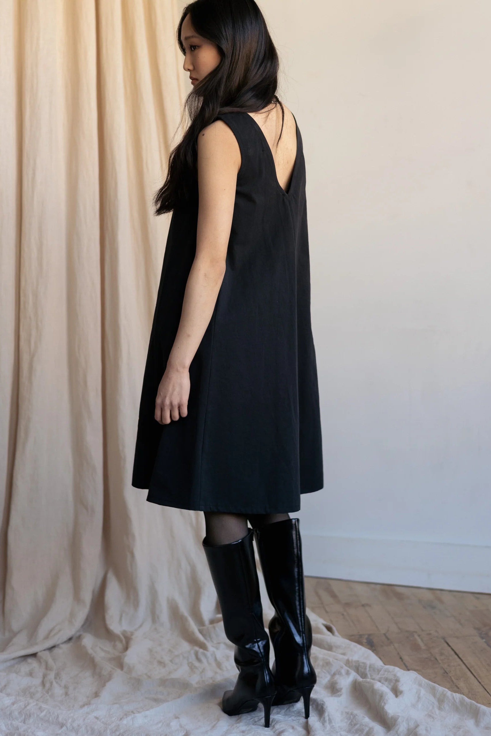 Side view of a woman wearing the Caro Dress by Lights of All in Black, a sleeveless knee-length trapeze dress with a round neck at the front and a V at the back. She is wearing it with knee-high black boots standing by a beige curtain