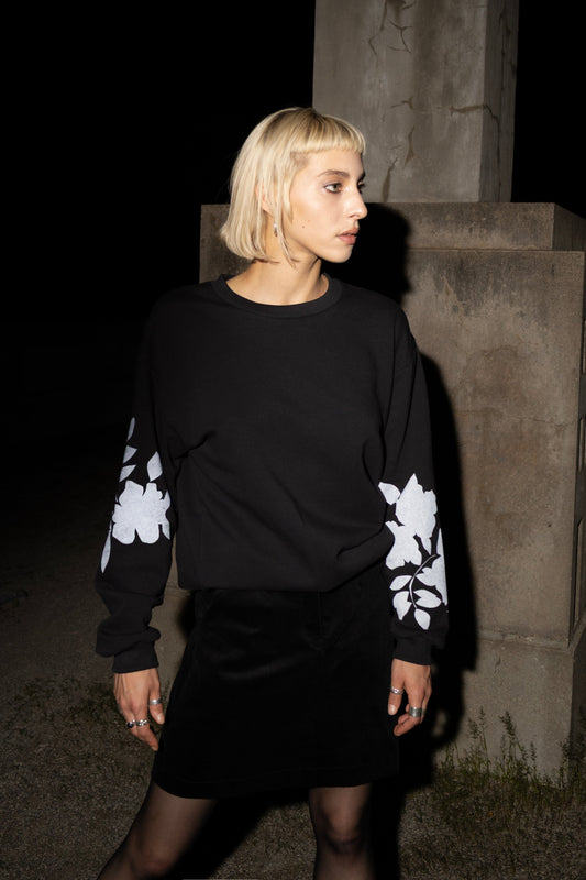 A woman wearing the Screen-Printed Crewneck Sweater by Eve Lavoie in Black with white floral patterns on the sleeves, paired with a black skirt, standing outdoors at night next to a concrete pillar.