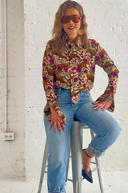 A woman wearing the Untamed Bloom Shirt by Dotty, a menswear-inspired blouse with a floral and animal print, classic button front details, and polished gold buttons. She is wearing it with jeans and sitting on a stool in front of a white brick wall. 