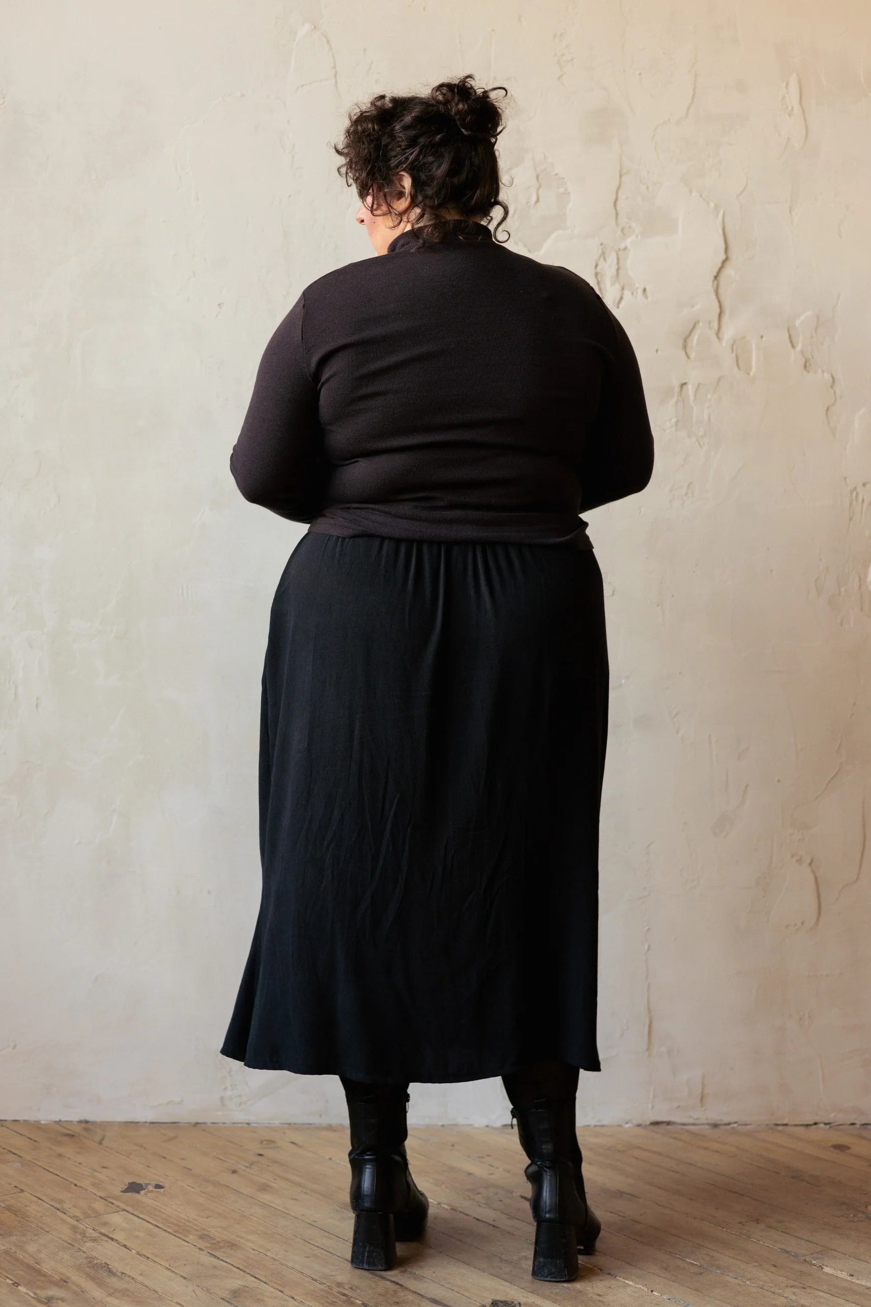 Back view of a woman wearing the Simone Skirt by Lights of All in Black, a sleek calf-length skirt with a flared hem and an elastic waist. She is wearing it with a brown turtleneck and standing by a beige wall. 