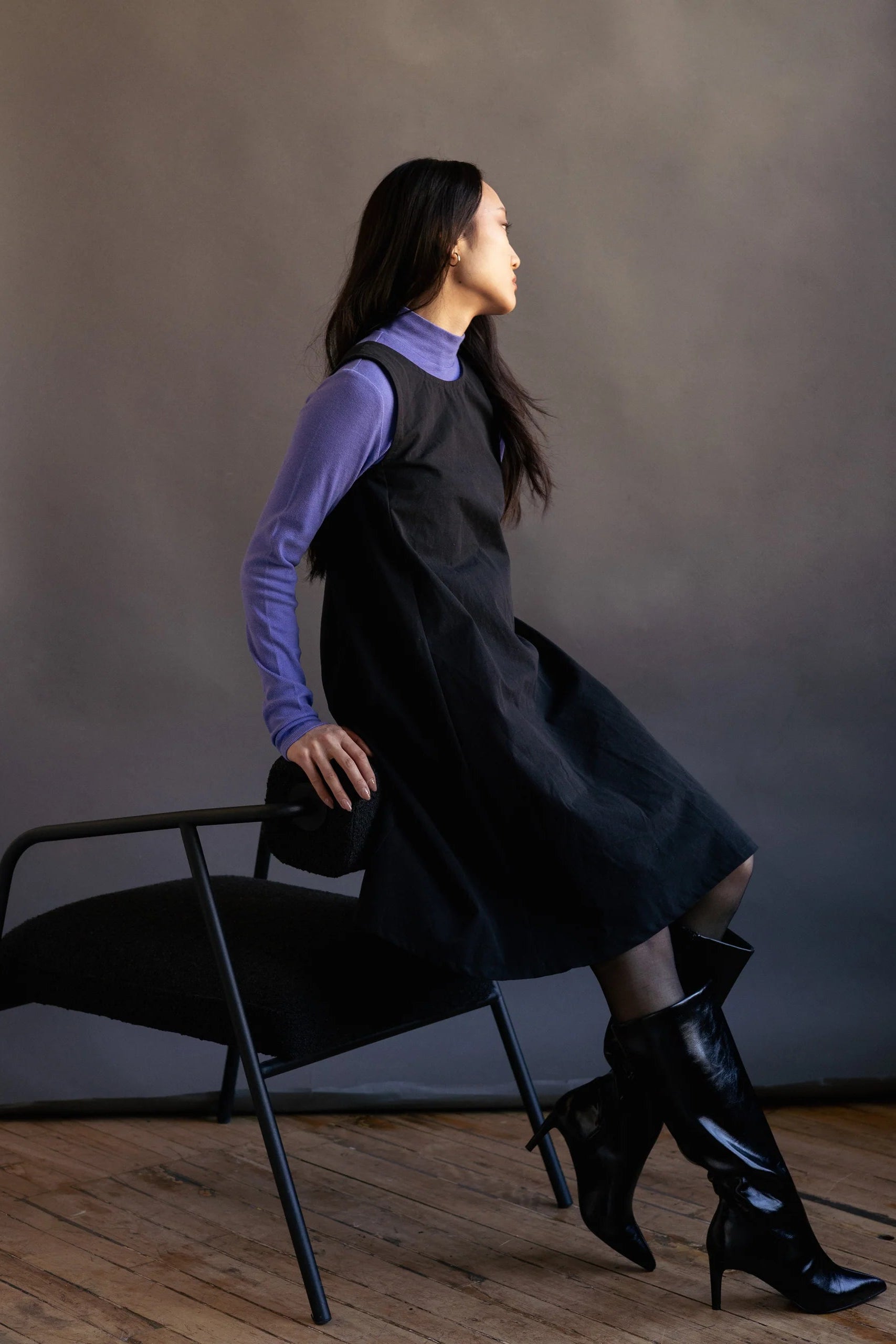 Side view of a woman wearing the Caro Dress by Lights of All in Black, a sleeveless knee-length trapeze dress with a round neck at the front and a V at the back. She is wearing it with a violet turtleneck and knee-high black boots and sitting on the arm of a chair in front of a dark background. 