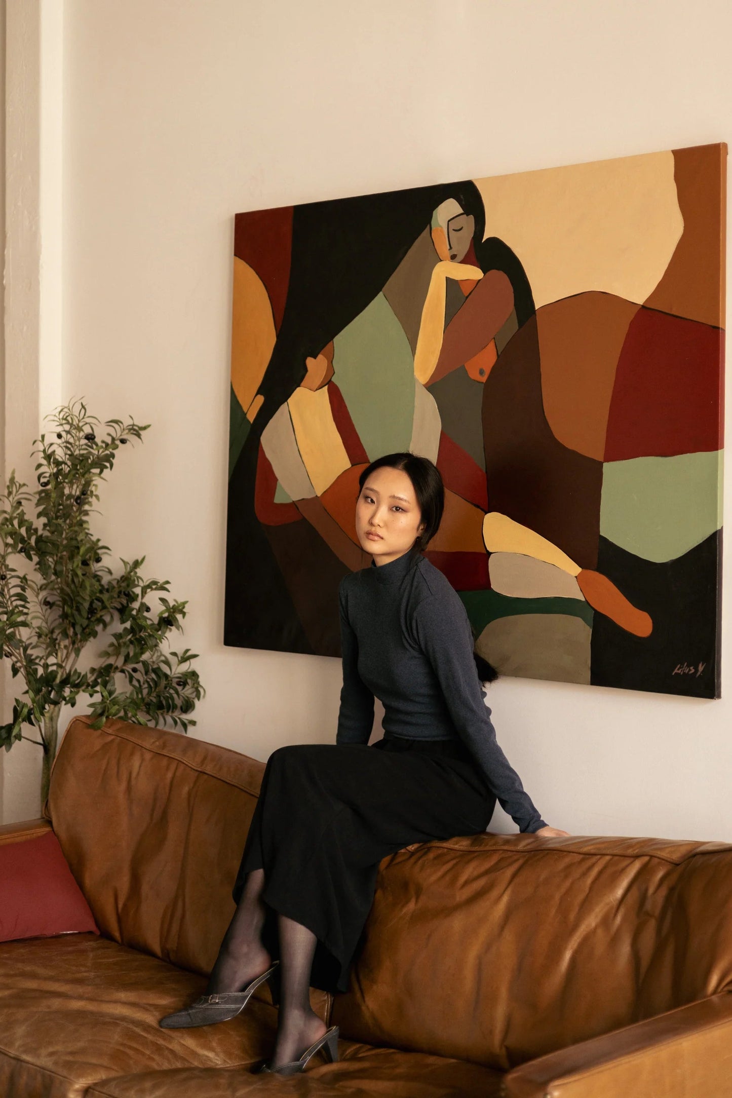 A woman wearing the Simone Skirt by Lights of All in Black, a sleek calf-length skirt with a flared hem and an elastic waist. She is wearing it with a grey turtleneck and sitting on a brown sofa in front of a large painting. 