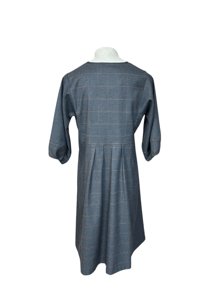 A back view of the Hanna Dress by Solomia in Grey is shown on a mannequin against a white background. This knee length dress has uniquely placed pleats and seams to create an architectural shape, with 3/4 length sleeves with structured cuffs. It has checked fabric on the front, back and cuffs, and solid grey fabric at the sides. 