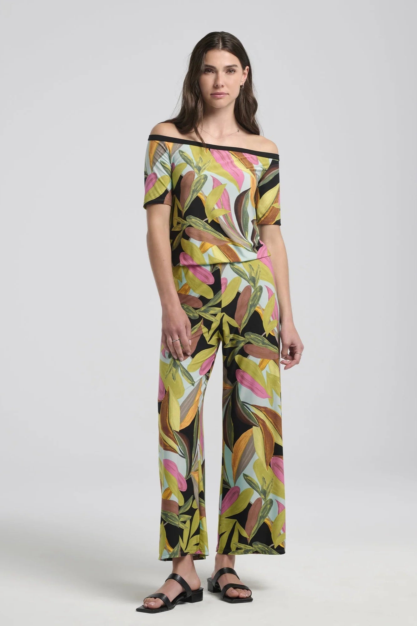 A woman wearing the Anthurium Jumpsuit by Kollontai in a Multicolour tropical print, with an off the shoulder neckline, bloused elastic waist, short sleeves and wide 7/8 length legs. She is standing in front of a grey background.l 