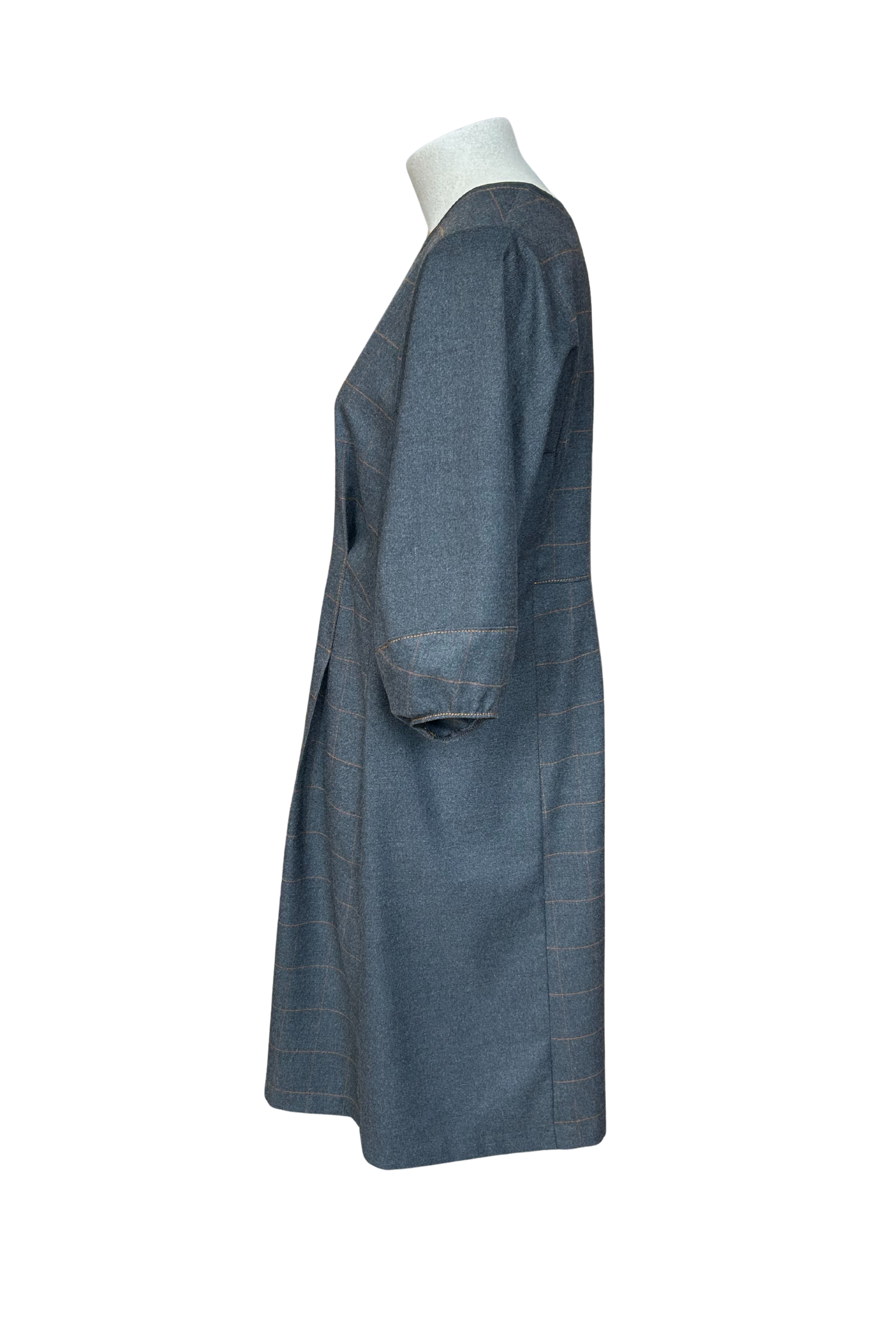 A side view of the Hanna Dress by Solomia in Grey is shown on a mannequin against a white background. This knee length dress has uniquely placed pleats and seams to create an architectural shape, with 3/4 length sleeves with structured cuffs. It has checked fabric on the front, back and cuffs, and solid grey fabric at the sides. 