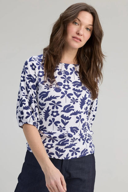 A woman wearing the Cadieux Top from message Factory in a floral Navy Print, with 3/4 sleeves with gathered cuffs, a wide neck and slim fit.  She is wearing it with dark pants and standing against a plain background. 