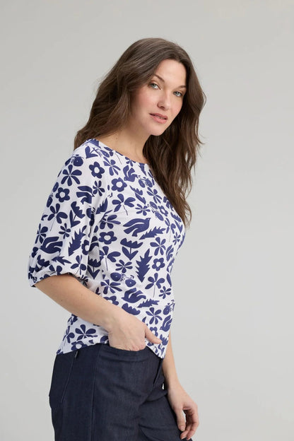 Side view of a woman wearing the Cadieux Top from Message Factory in a floral Navy Print, with 3/4 sleeves with gathered cuffs, a wide neck and slim fit.  She is wearing it with dark pants and standing against a plain background. 