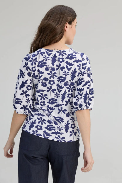 Back view of a woman wearing the Cadieux Top from message Factory in a floral Navy Print, with 3/4 sleeves with gathered cuffs, a wide neck and slim fit.  She is wearing it with dark pants and standing against a plain background. 
