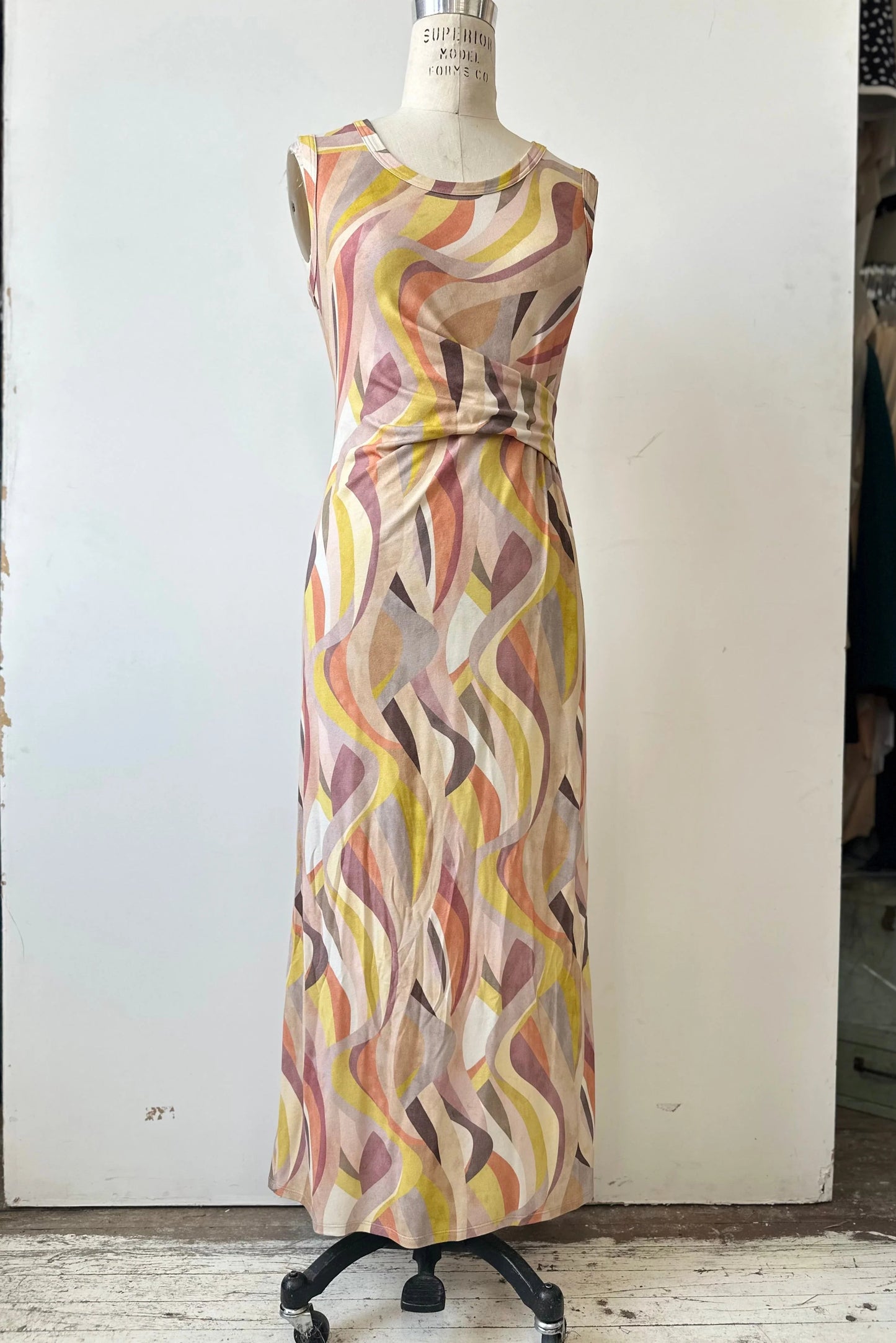 The Serina Dress by Kollontai in Peach print, a long, slim cut sleeveless dress with a soft pleat at the waist, is shown on a mannequin in front of a white background. 