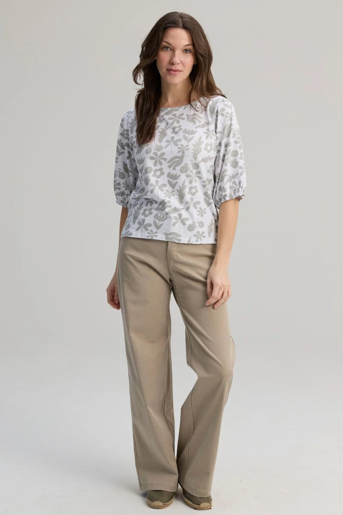 A woman wearing the Cadieux Top from Message Factory in a floral White Print, with 3/4 sleeves with gathered cuffs, a wide neck and slim fit.  She is wearing it with tan pants and standing against a plain background. 