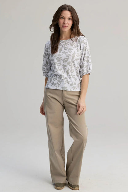 A woman wearing the Cadieux Top from Message Factory in a floral White Print, with 3/4 sleeves with gathered cuffs, a wide neck and slim fit.  She is wearing it with tan pants and standing against a plain background. 