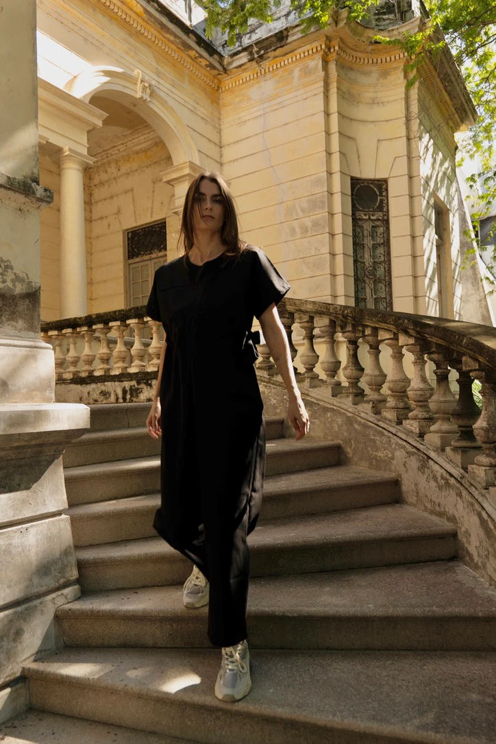 A woman wearing the Oslo Jumpsuit by MAS in Pepper, with round neck, short sleeves, adjustable waist tie, and wide legs. She is walking down a set out of outdoor stairs. 