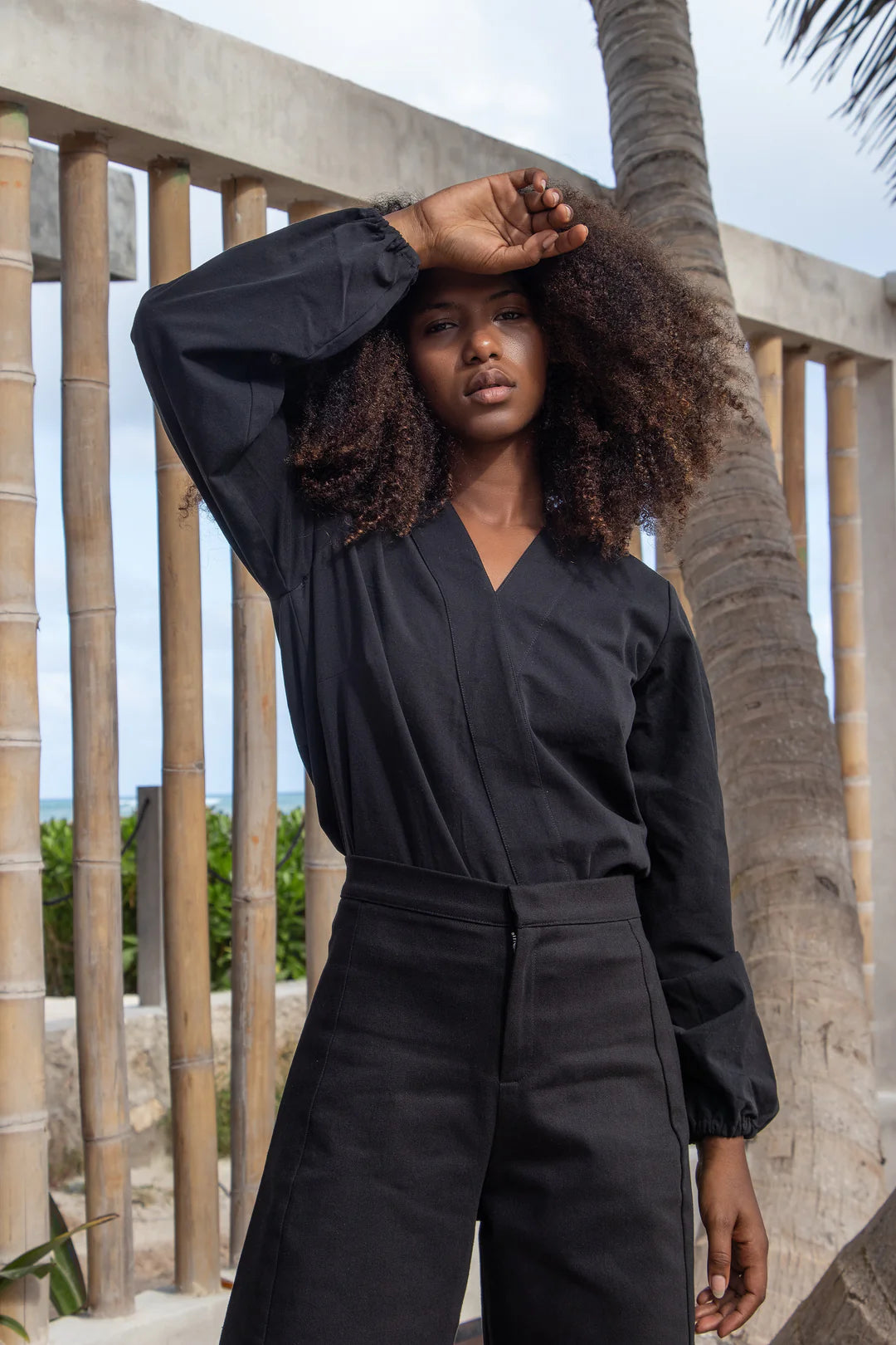 A woman wearing the Vienna Pants by MAS in Pepper, with a wide leg, side seams that sit slightly to the front, and two back pockets, with a black blouse, standing in front of a wooden wall. 