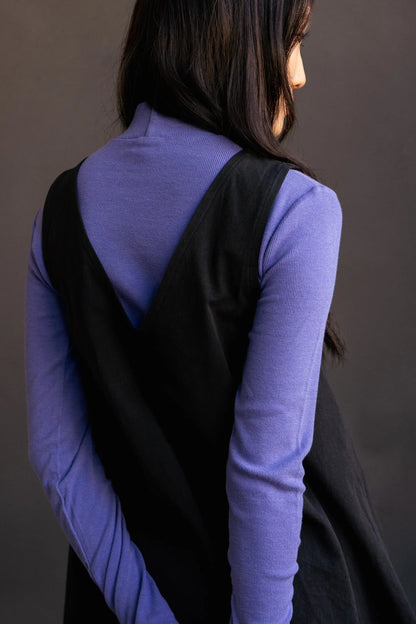 Back close up of view of a woman wearing the Caro Dress by Lights of All in Black, a sleeveless knee-length trapeze dress with a round neck at the front and a V at the back. She is wearing it over a violet turtleneck and standing in front of a grey background. 