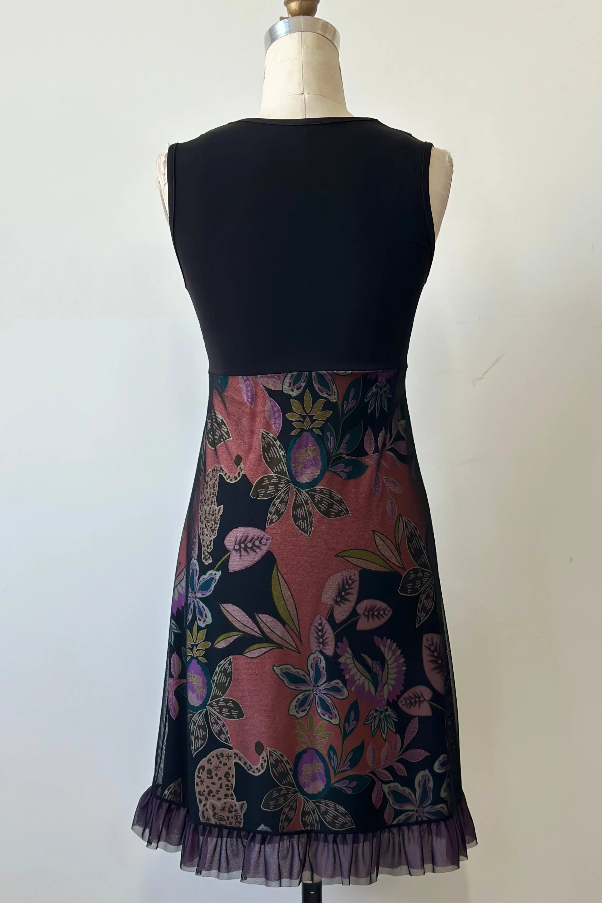 The back of the Liseron Dress by Kollontai is shown on a mannequin against a grey background. It's a sleeveless dress with a faux-wrap neckline, empire waist, knee-length a-line skirt. The bodice is solid black, while the skirt features a cheetah/floral print under a mesh overlay. 