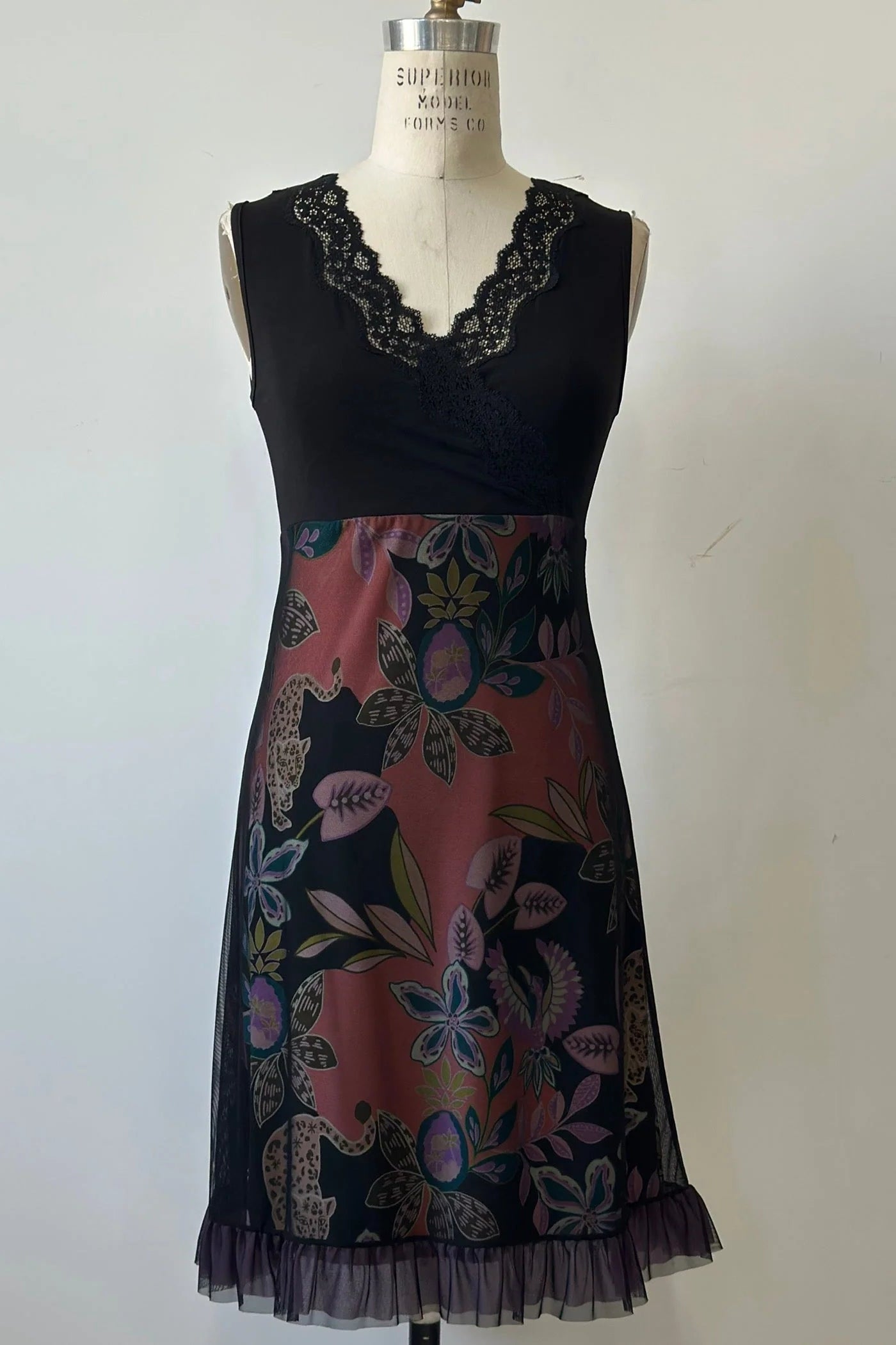 The Liseron Dress by Kollontai is shown on a mannequin against a grey background. It's a sleeveless dress with a faux-wrap neckline, empire waist, knee-length a-line skirt. The bodice is solid black, while the skirt features a cheetah/floral print under a mesh overlay. 