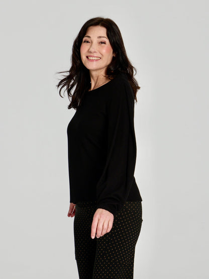 Side view of A woman wearing the Virginia Top by Kollontai in Black, a corduroy-textured top with inverted pleats on the long sleeves, a slim fit and a round neck. She is wearing it with the Susan pants in Black and standing in front of a grey background. 