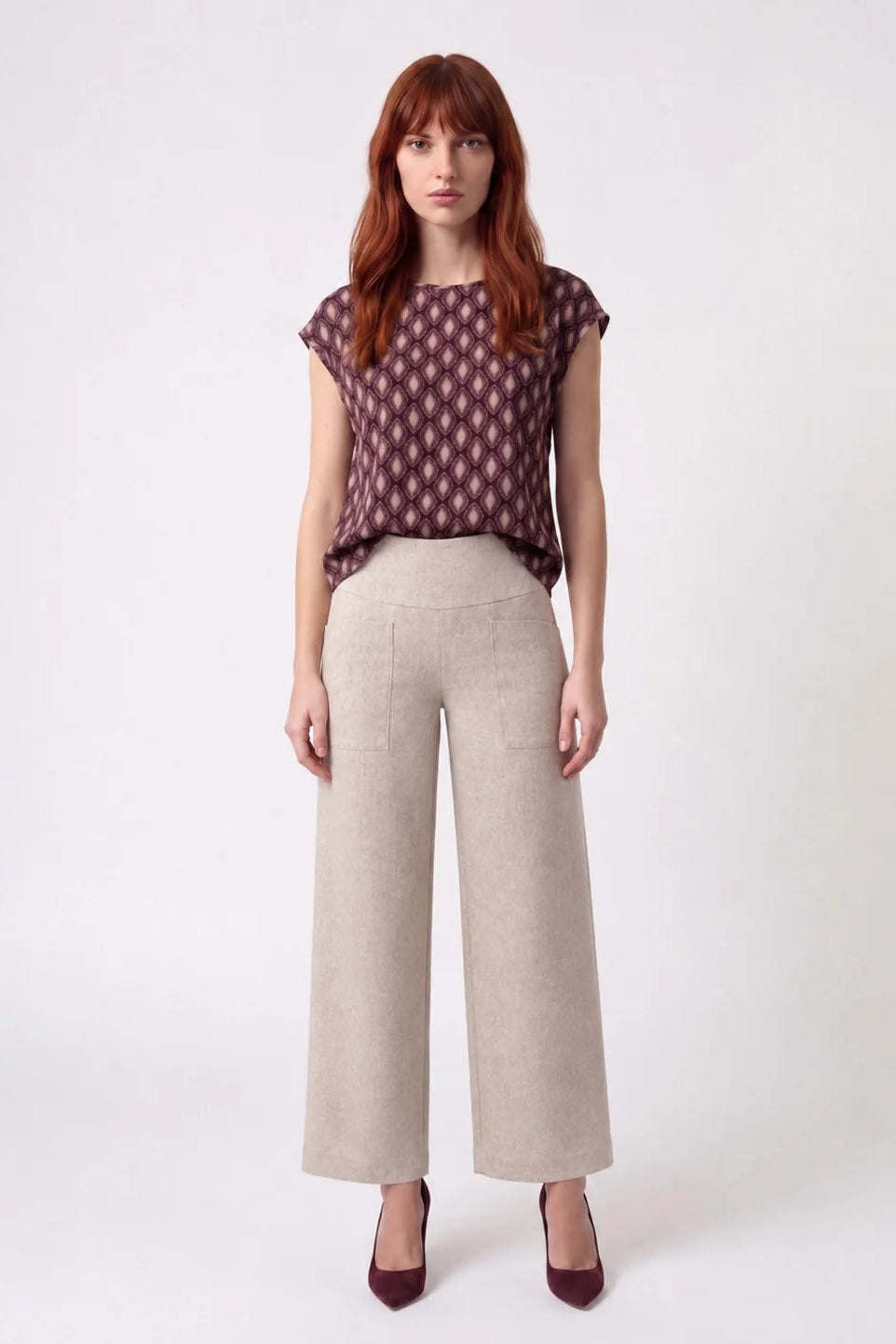 A woman wearing the Fanie Shirt by Kollontai, a relaxed fitting top with cape sleeves and a hi-low hem in a pink and plum geometric print. She is wearing it with cream pants and standing against a white background. 