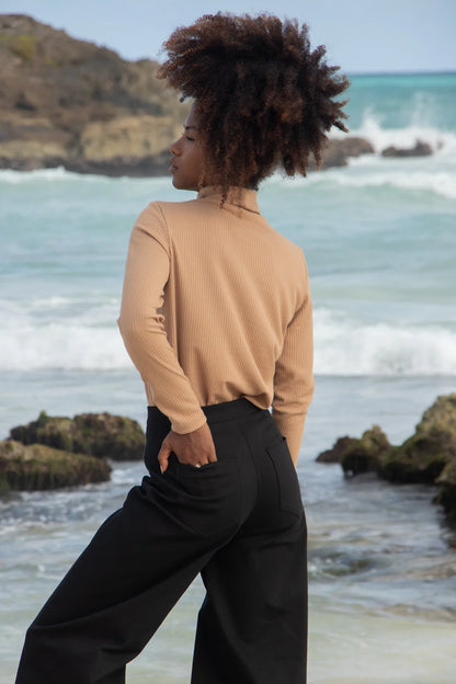 A woman wearing the Vienna Pants by MAS in Pepper with a wide leg, side seams that sit slightly to the front, and two back pockets, with tan shirt. She is standing in front of the ocean. 