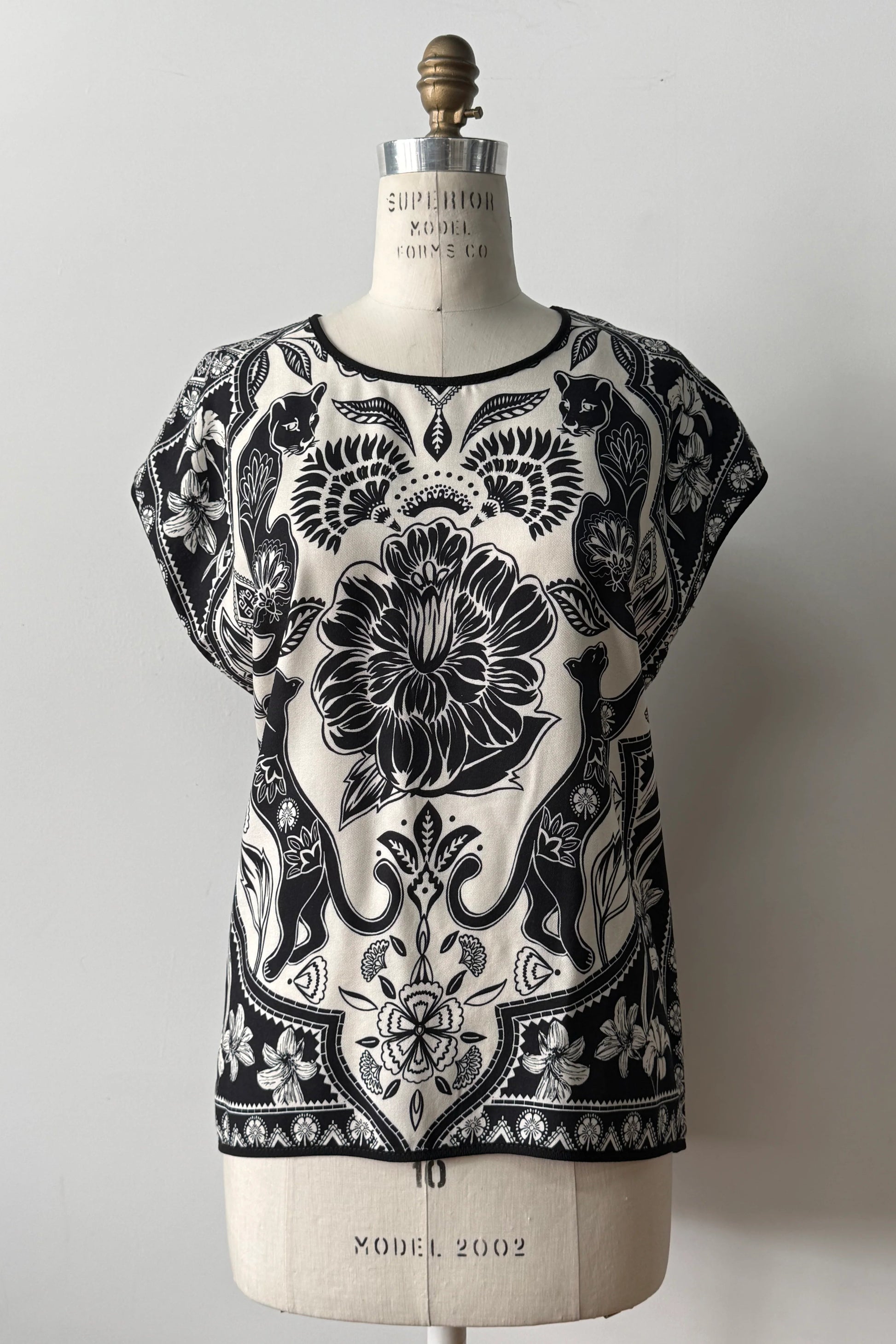 The Malo T-shirt from Kollontai, featuring a floral and panther print in black-on-ivory on the front, cap sleeves, and a solid black back, is shown on a mannequin in front of a grey background 