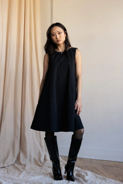 A woman wearing the Caro Dress by Lights of All in Black, a sleeveless knee-length trapeze dress with a round neck at the front and a V at the back. She is wearing it with knee-high black boots standing against a beige curtain.