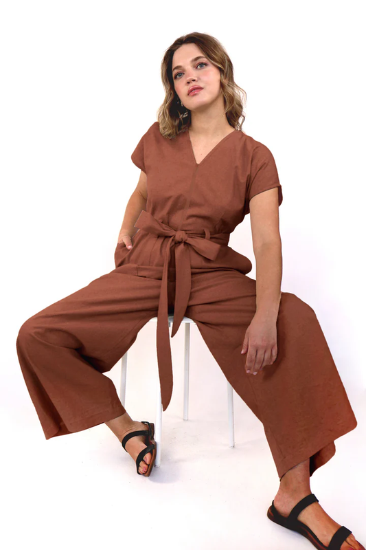 A woman wearing the Alexandra Long Jumpsuit by Korelli in Rust, seated on a chair in front of a white background