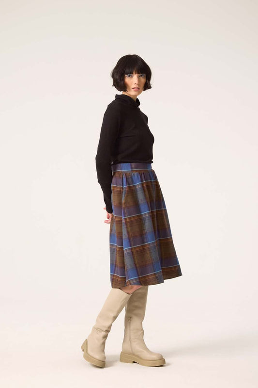 Side view of a woman wearing the Alisma Skirt by Cokluch in Plaid Blue with a black turtleneck, standing in front of a white background 