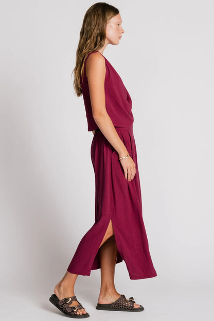 Side view of a woman wearing the Barbet Skirt by Allison Wonderland in Pink, a maxi dress with side slits, front pleats, elastic at the back waist and a side button detail at the front. She is wearing it with a matching tank and standing in front of a white background. 