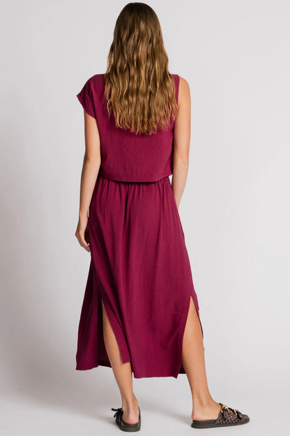 Back view of a woman wearing the Barbet Skirt by Allison Wonderland in Pink, a maxi dress with side slits, front pleats, elastic at the back waist and a side button detail at the front. She is wearing it with a matching tank and standing in front of a white background. 