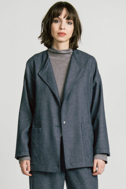 3/4 view of a woman wearing the Bootes Blazer by Allison Wonderland in Denim, a slouchy collarless blazer with a single waist button and a shoulder button, and drop sleeves, with large patch pockets. She is wearing it with the  Octans turtleneck in Grey and the matching Lynx Pants and standing against a light grey studio background.