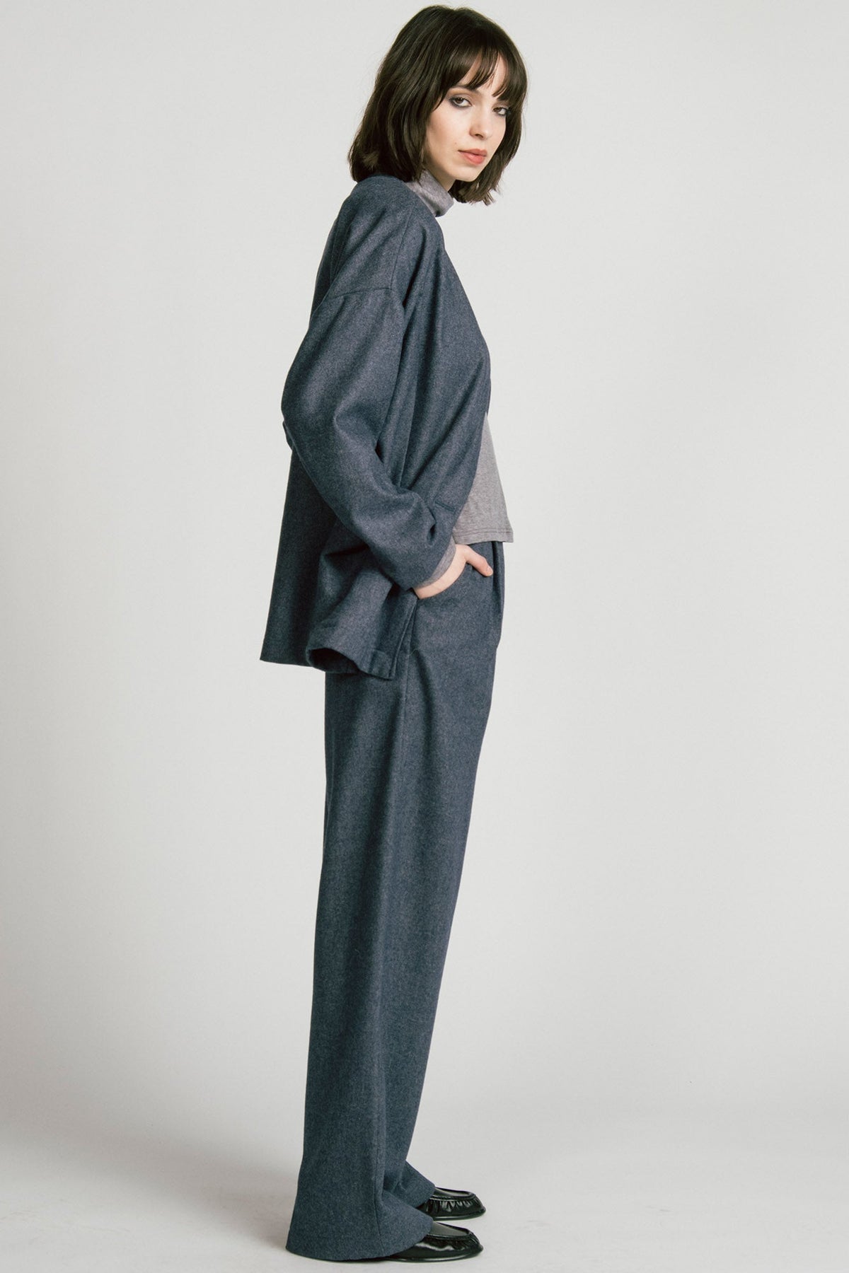 Side view of a woman wearing the Bootes Blazer by Allison Wonderland in Denim, a slouchy collarless blazer with  a single waist button and a shoulder button, and drop sleeves, with large patch pockets. She is wearing it with the  Octans turtleneck in Grey and the matching Lynx Pants and standing against a light grey studio background.