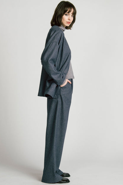 Side view of a woman wearing the Bootes Blazer by Allison Wonderland in Denim, a slouchy collarless blazer with  a single waist button and a shoulder button, and drop sleeves, with large patch pockets. She is wearing it with the  Octans turtleneck in Grey and the matching Lynx Pants and standing against a light grey studio background.