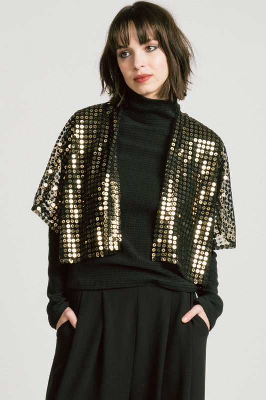 3/4 view of a  woman wearing the Corvus Shrug by Allison Wonderland in Sequins, a sheer black bolero with golden sequins. She is wearing it over a black turtleneck and black pants and standing in front of a white background. 