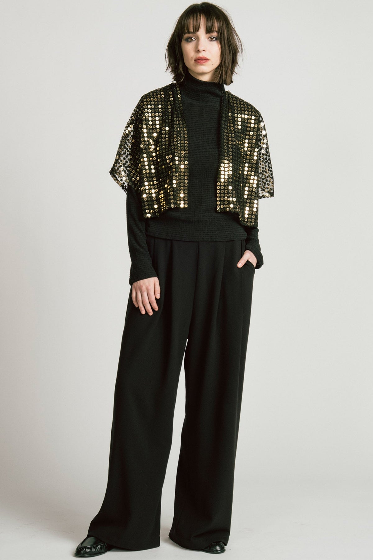 A woman wearing the Corvus Shrug by Allison Wonderland in Sequins, a sheer black bolero with golden sequins. She is wearing it over a black turtleneck and black pants and standing in front of a white background. 