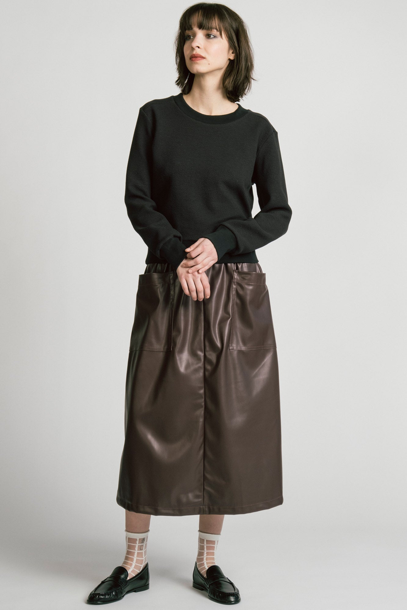 A woman wearing the Crux Sweatshirt by Allison Wonderland in Black Texture. she is wearing it with a brown pleather skirt and standing in front of a white background. 