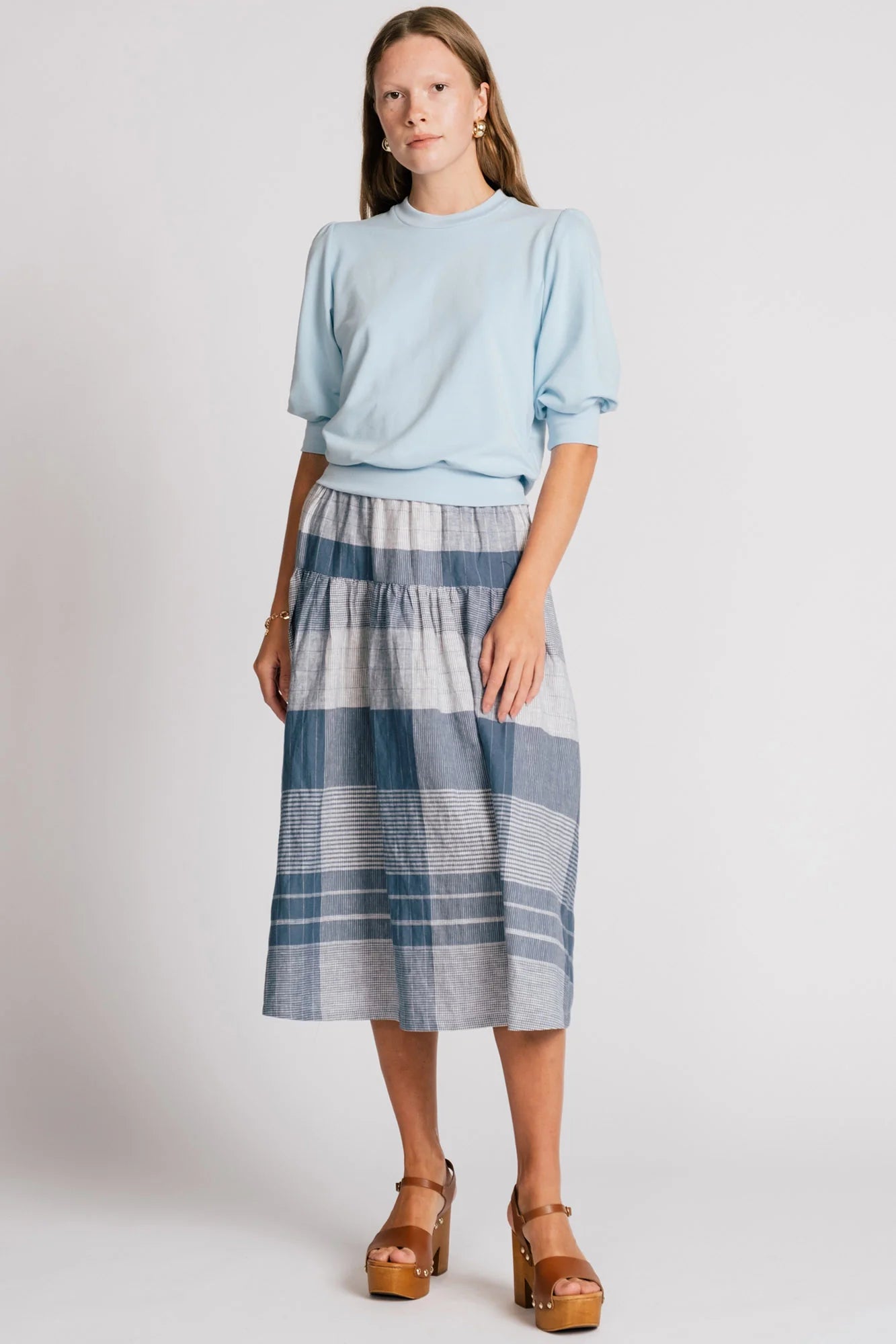 A woman wearing the Finch Sweatshirt by Allison Wonderland in Baby Blue, a cropped sweatshirt with elbow-length puffed sleeves and ribbing at the neck and waistband. She is wearing it with a blue plaid skirt and standing in front of a white background.