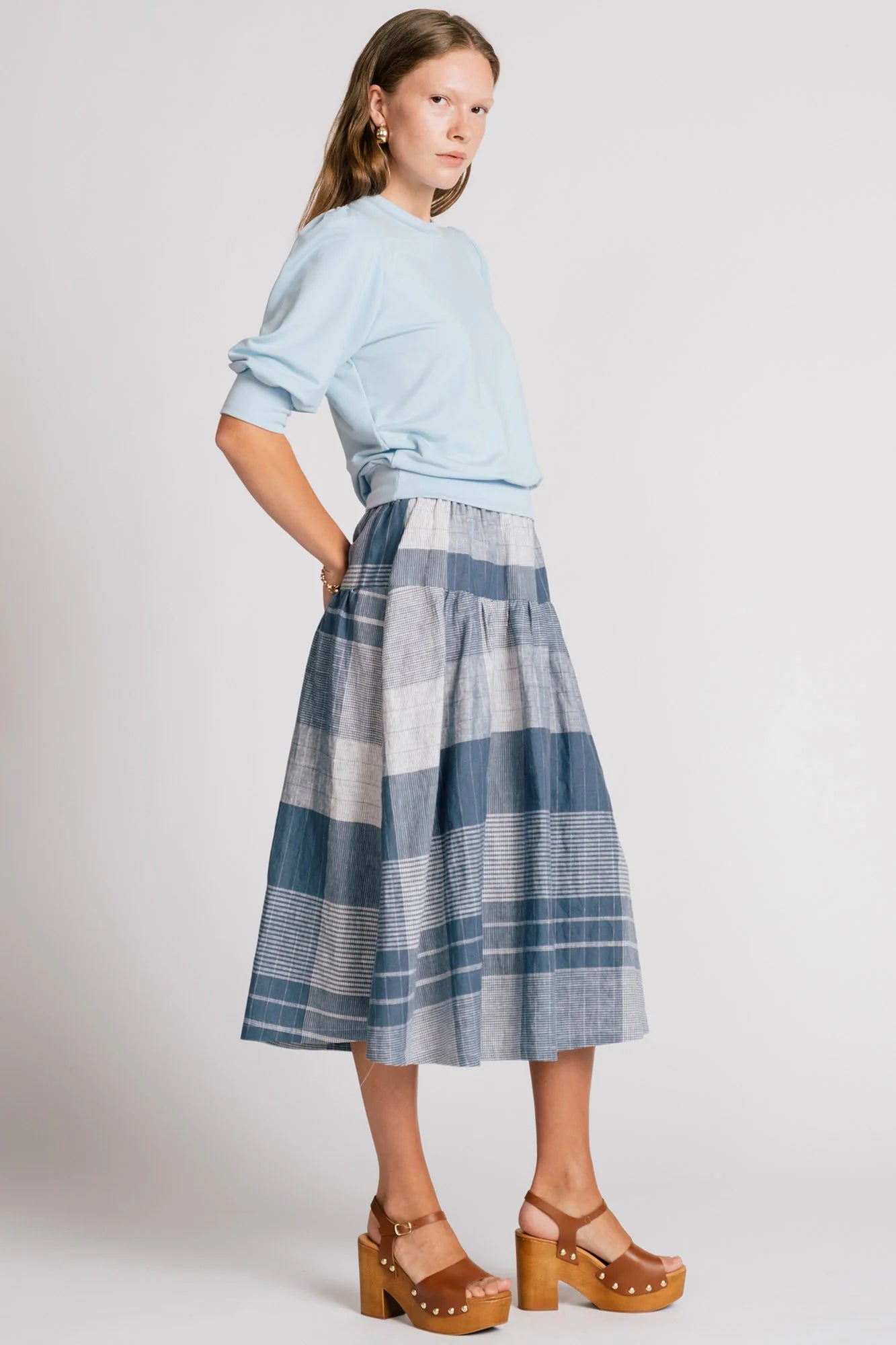 Side view of a woman wearing the Finch Sweatshirt by Allison Wonderland in Baby Blue, a cropped sweatshirt with elbow-length puffed sleeves and ribbing at the neck and waistband. She is wearing it with a blue plaid skirt and standing in front of a white background.