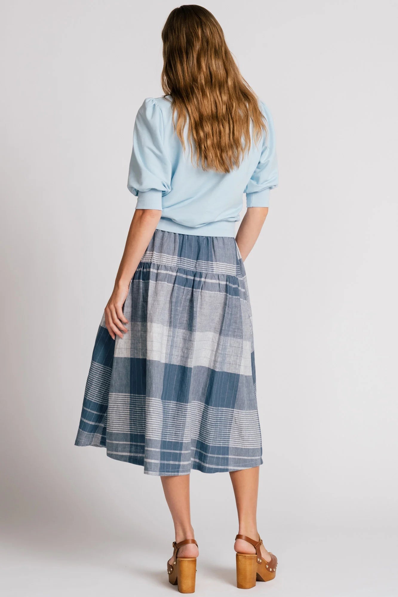 Back view of a woman wearing the Finch Sweatshirt by Allison Wonderland in Baby Blue, a cropped sweatshirt with elbow-length puffed sleeves and ribbing at the neck and waistband. She is wearing it with a blue plaid skirt and standing in front of a white background.