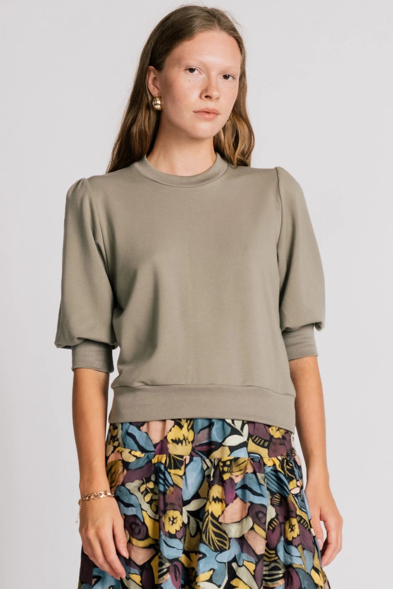 A woman wearing the Finch Sweatshirt by Allison Wonderland in Eucalyptus, a cropped sweatshirt with elbow-length puffed sleeves and ribbing at the neck and waistband. She is wearing it with a patterned skirt and standing in front of a white background.