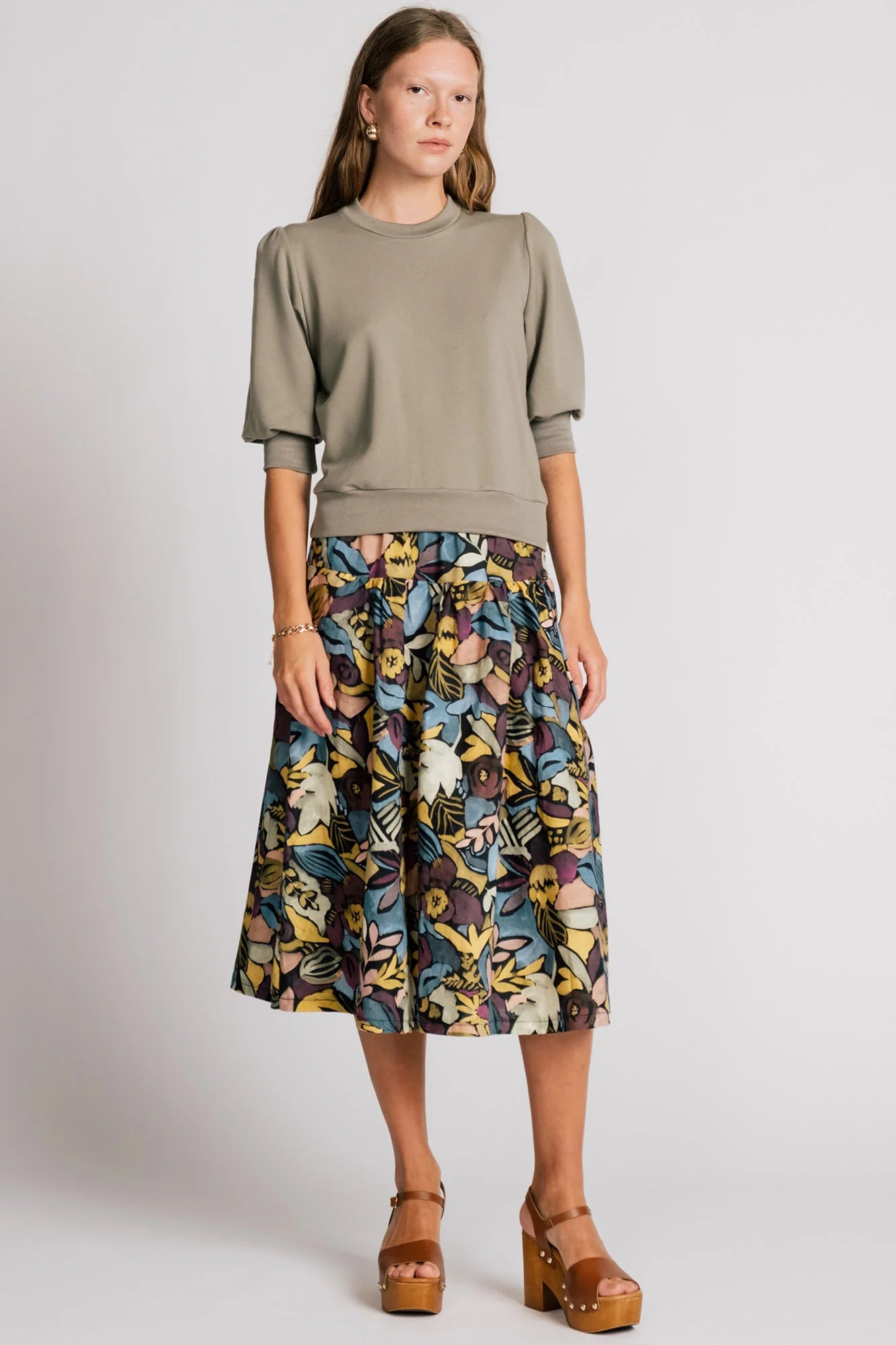 A woman wearing the Finch Sweatshirt by Allison Wonderland in Eucalyptus, a cropped sweatshirt with elbow-length puffed sleeves and ribbing at the neck and waistband. She is wearing it with a patterned skirt and standing in front of a white background.