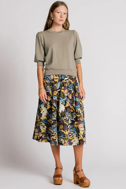 A woman wearing the Finch Sweatshirt by Allison Wonderland in Eucalyptus, a cropped sweatshirt with elbow-length puffed sleeves and ribbing at the neck and waistband. She is wearing it with a patterned skirt and standing in front of a white background.