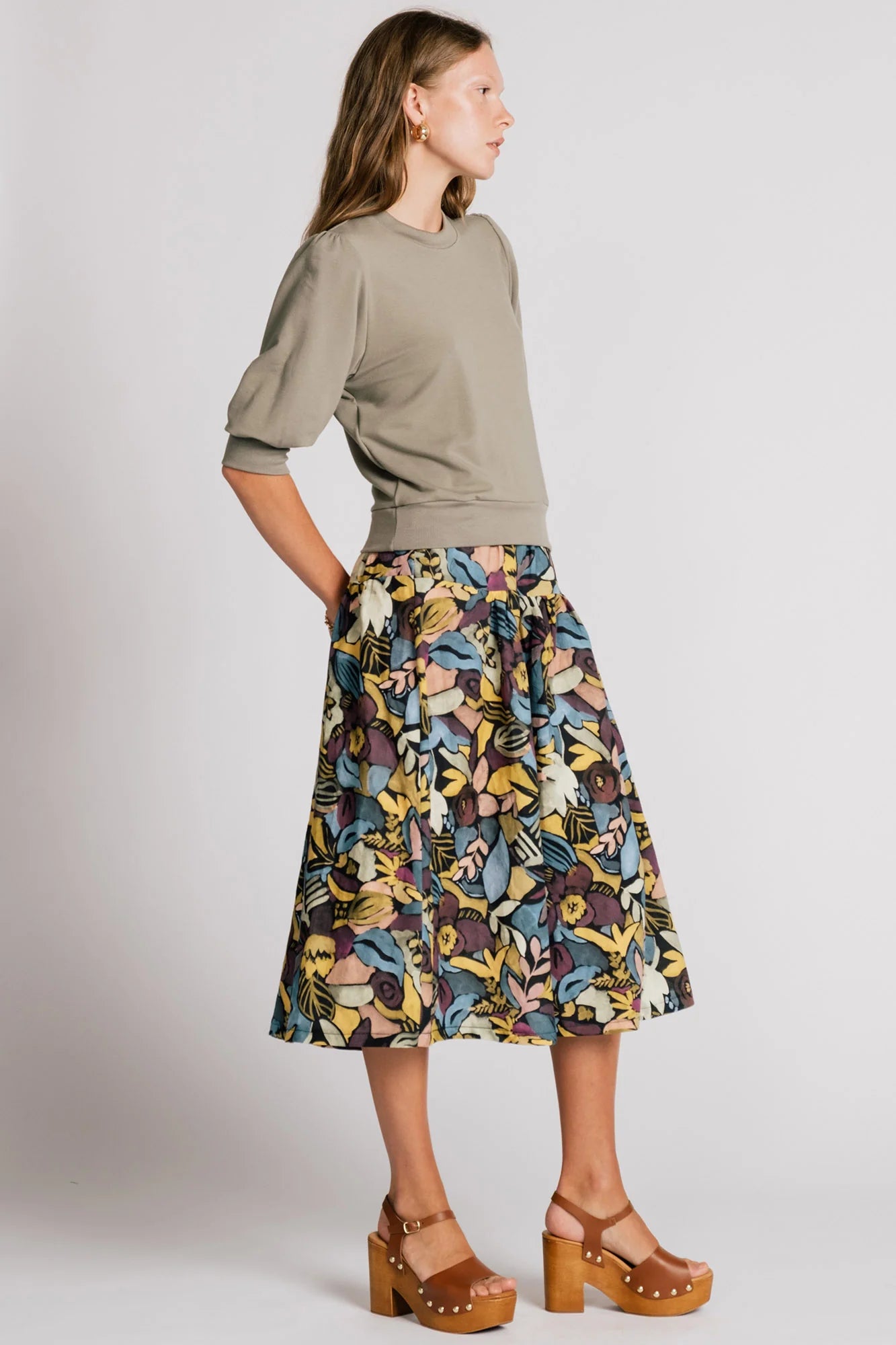 Side view of a woman wearing the Finch Sweatshirt by Allison Wonderland in Eucalyptus, a cropped sweatshirt with elbow-length puffed sleeves and ribbing at the neck and waistband. She is wearing it with a patterned skirt and standing in front of a white background.