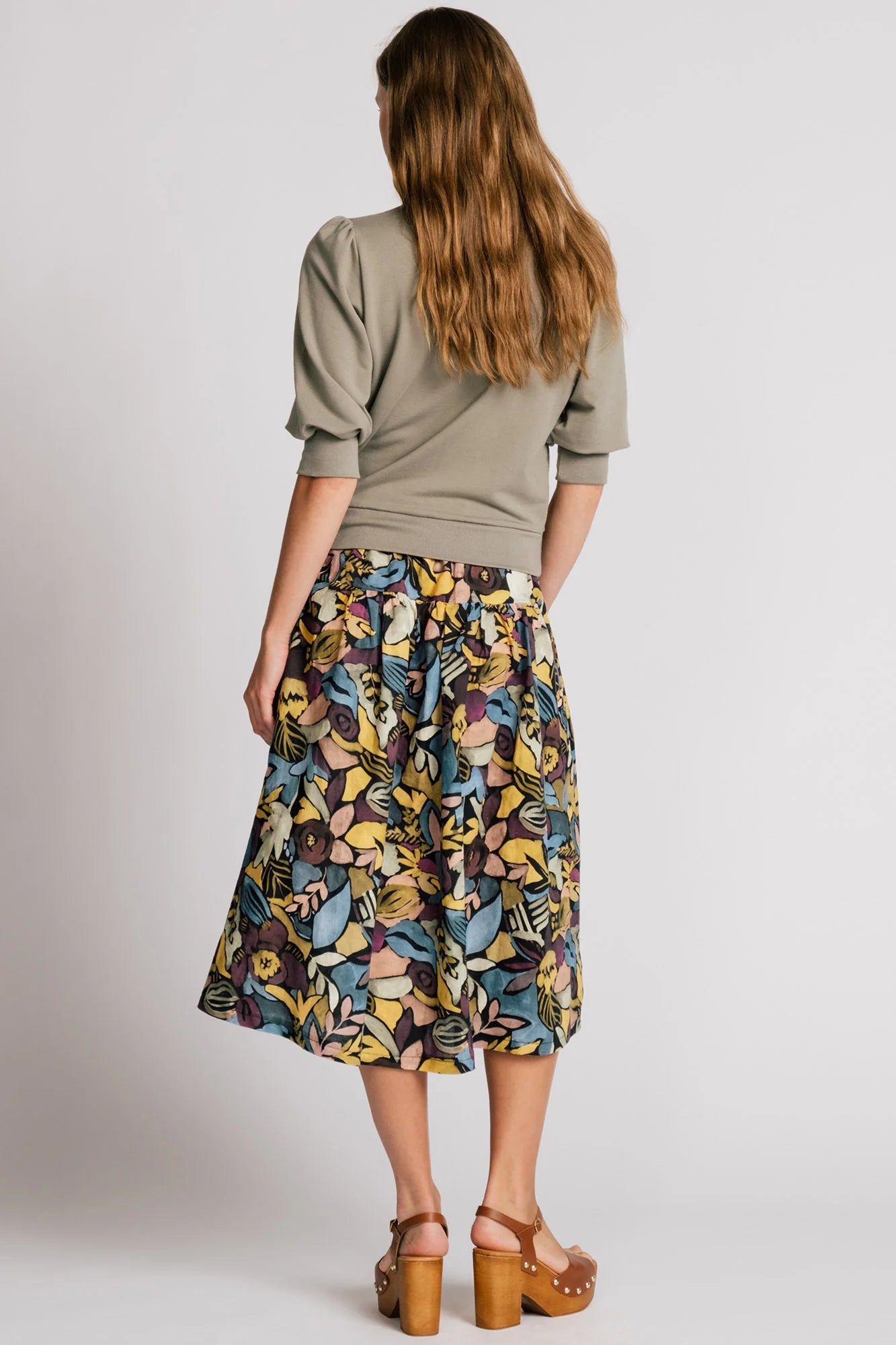 Back view of a woman wearing the Finch Sweatshirt by Allison Wonderland in Eucalyptus, a cropped sweatshirt with elbow-length puffed sleeves and ribbing at the neck and waistband. She is wearing it with a patterned skirt and standing in front of a white background.