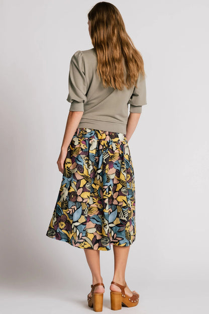 Back view of a woman wearing the Finch Sweatshirt by Allison Wonderland in Eucalyptus, a cropped sweatshirt with elbow-length puffed sleeves and ribbing at the neck and waistband. She is wearing it with a patterned skirt and standing in front of a white background.
