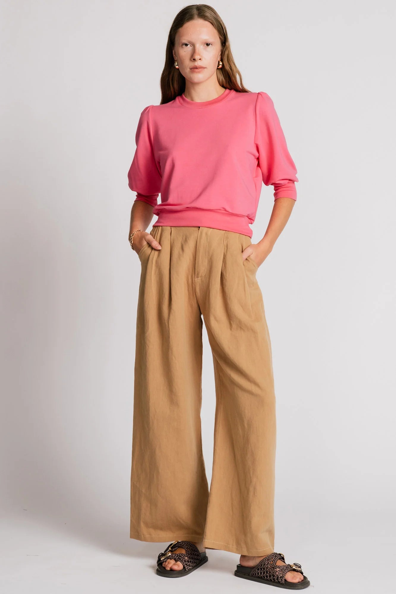 A woman wearing the Finch Sweatshirt by Allison Wonderland in Pink, a cropped sweatshirt with elbow-length puffed sleeves and ribbing at the neck and waistband. She is wearing it with beige pants and standing in front of a white background.