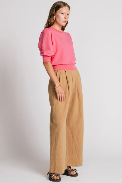 Side view of a woman wearing the Finch Sweatshirt by Allison Wonderland in Pink, a cropped sweatshirt with elbow-length puffed sleeves and ribbing at the neck and waistband. She is wearing it with beige pants and standing in front of a white background.