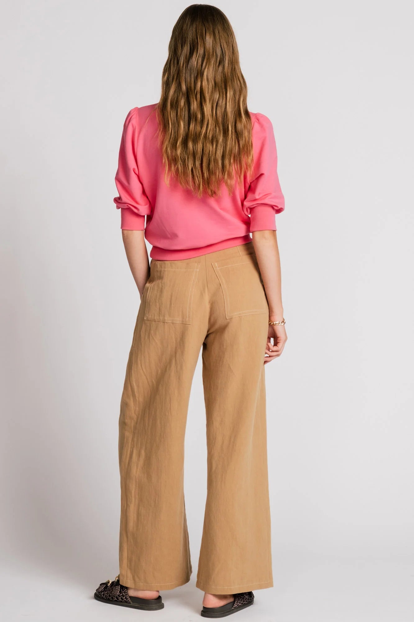 Back view of a woman wearing the Finch Sweatshirt by Allison Wonderland in Pink, a cropped sweatshirt with elbow-length puffed sleeves and ribbing at the neck and waistband. She is wearing it with beige pants and standing in front of a white background.