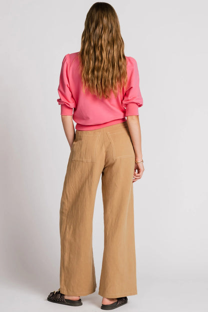 Back view of a woman wearing the Finch Sweatshirt by Allison Wonderland in Pink, a cropped sweatshirt with elbow-length puffed sleeves and ribbing at the neck and waistband. She is wearing it with beige pants and standing in front of a white background.